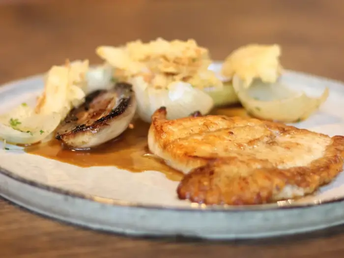 En tallerken med et kogt kyllingebryst, ristede løg og noget, der ligner kartoffelchips, serveret med brun sauce.
