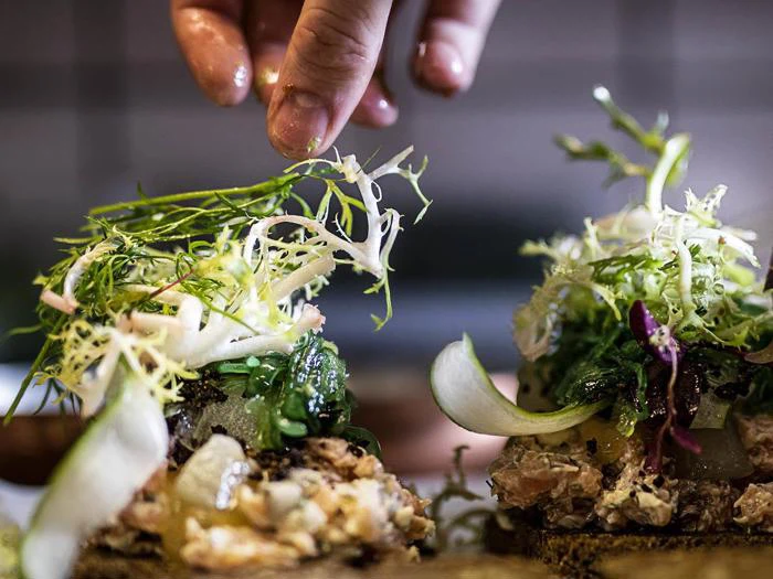 En hånd drysser frisk grønt på smørrebrød med forskellige grøntsager og pålæg og viser en detaljeret præsentation.