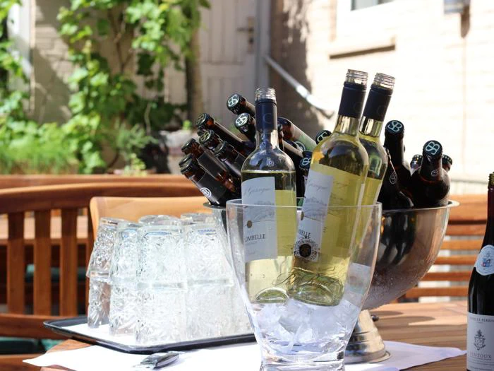 Flasker med hvidvin og øl står på køl i isspande på et udendørs træbord, og klare glas står i nærheden i sollyset.