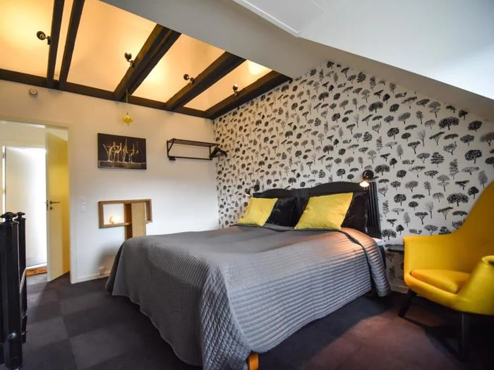 Et hyggeligt soveværelse med dobbeltseng, gule puder, mønstret tapet, synlige loftsbjælker, en gul stol og varm belysning.