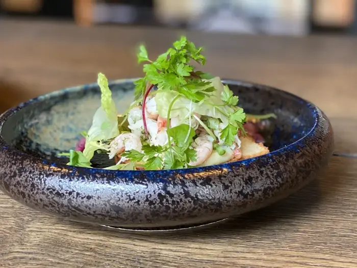 På et træbord står en rustik keramikskål fyldt med en frisk salat, toppet med bladgrønt og krydderurter.