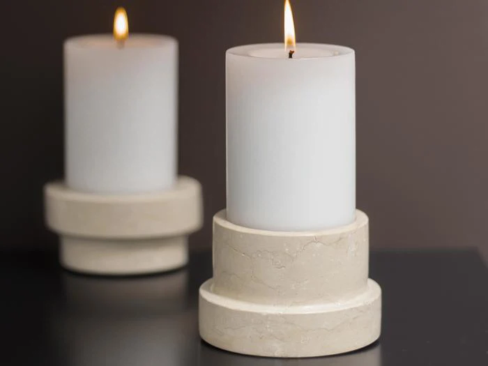 To hvide bloklys med flammer, hver placeret på en rund beige marmorholder, vises på en mørk overflade med en sløret baggrund.