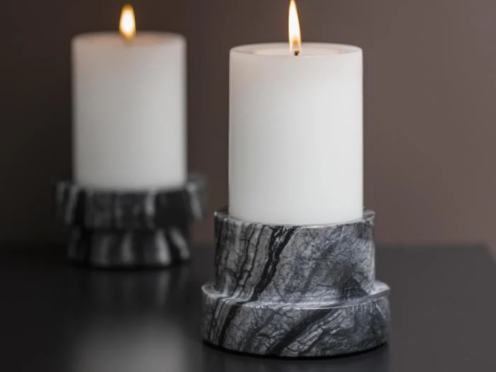 Et tændt hvidt bloklys i en sort og grå marmoreret holder står på en mørk overflade med et andet lignende lys sløret i baggrunden.