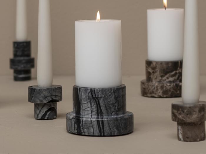 Et hvidt bloklys brænder i en mørk marmorholder, omgivet af utændte hvide bloklys i forskellige marmorholdere på en beige overflade.