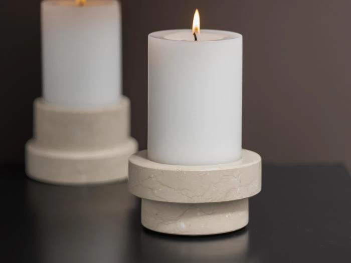 Et hvidt bloklys med en glat stenholder er tændt på en mørk overflade med et andet utændt lys i baggrunden.