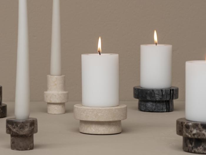 Flere hvide lys, både bloklys og fyrfadslys, er placeret i stenholdere i forskellige farver på en neutral overflade; tre lys er tændt.