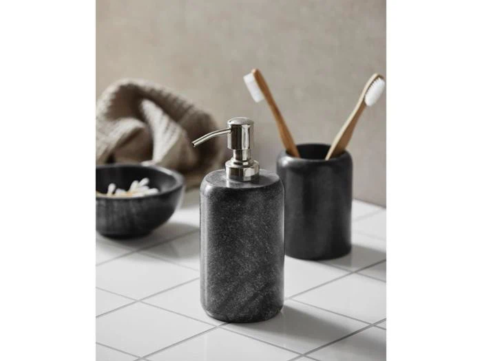 En sort sæbedispenser står på en hvid flisebelagt overflade ved siden af en matchende kop med to bambustandbørster og en lille skål med vatpinde.