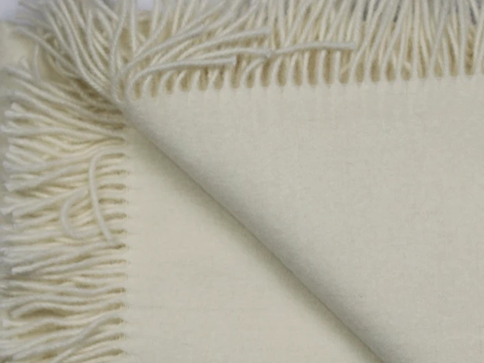 Nærbillede af et foldet cremefarvet uldtæppe med frynsede kanter, der viser blød tekstur og sirligt arrangerede kvaster.