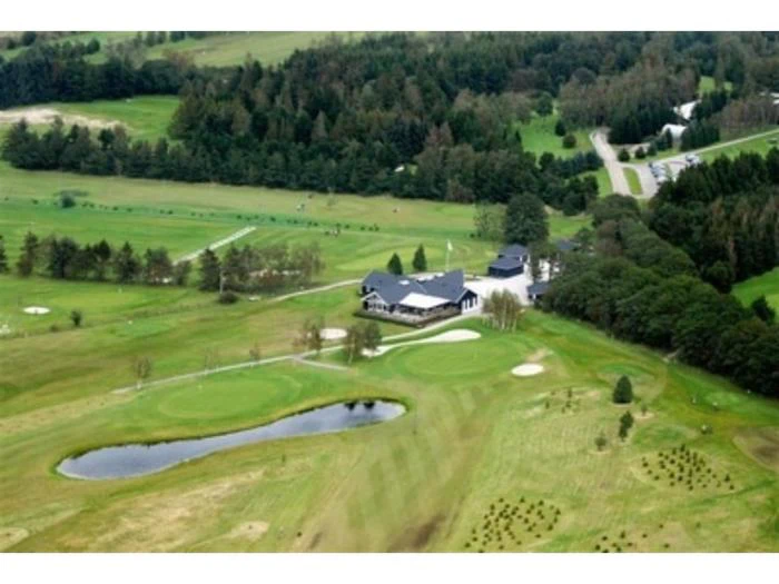Luftfoto af en golfbane med et klubhus omgivet af træer, græsklædte fairways, sandbunkers og en lille dam.