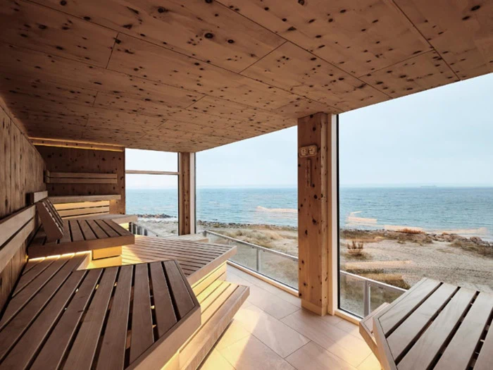 En moderne træsauna med brede bænke har udsigt til strand og hav gennem store gulv-til-loft-vinduer.