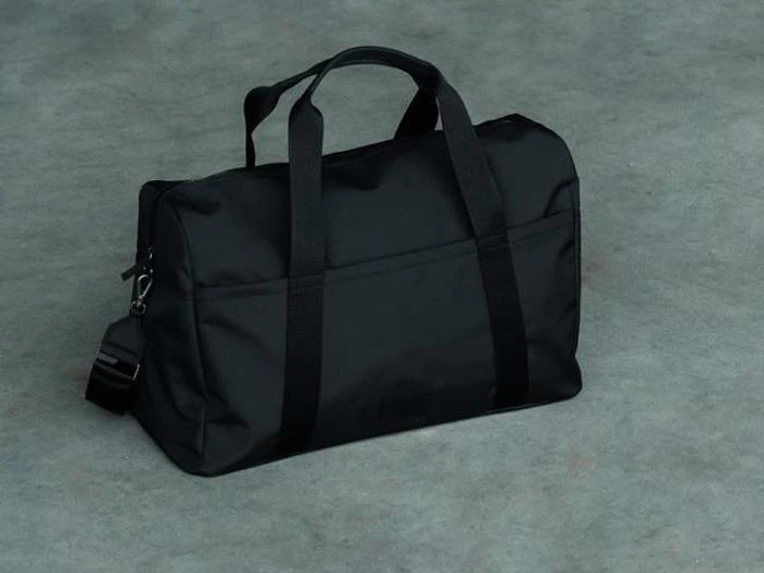 En sort duffelbag med håndtag og skulderrem ligger på en grå betonflade.