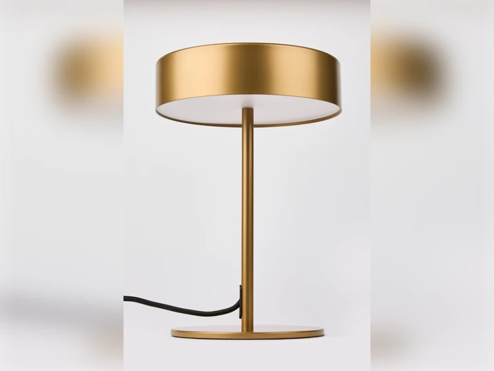 En moderne bordlampe i guld med en rund fod, et slankt stativ og en bred, cylindrisk lampeskærm, fotograferet på en ensfarvet hvid baggrund.