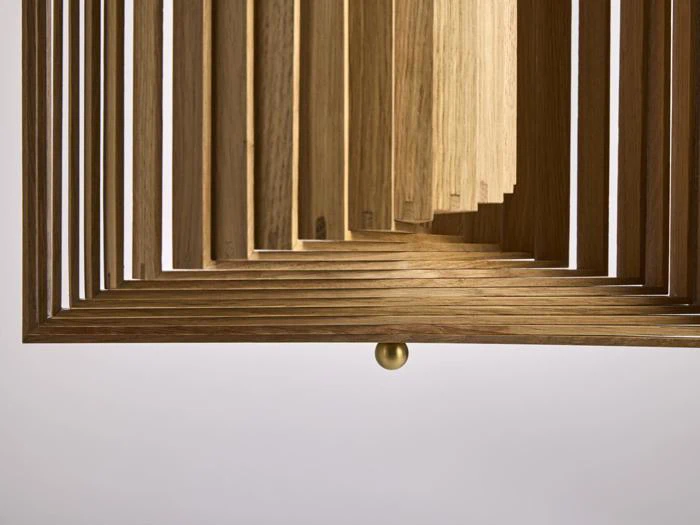 Nærbillede af en geometrisk træstruktur med lagdelte rektangulære lameller og en lille rund guldknap i bunden på en almindelig baggrund.