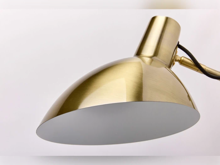 Nærbillede af en moderne skrivebordslampe i messing med kuppelformet skærm og sort ledning mod en almindelig lys baggrund.