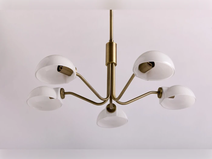En moderne lysekrone i messing med fem buede arme, der hver holder en hvid, kuppelformet lampeskærm, ophængt mod en almindelig lys baggrund.