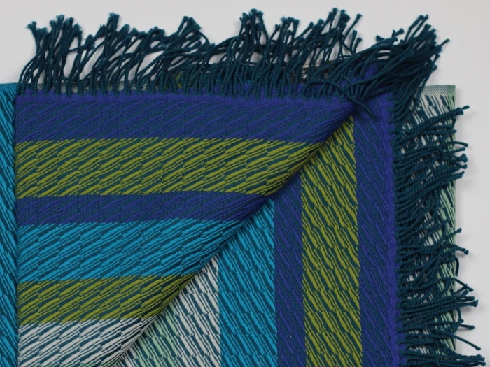 Et vævet stof med blå, grønne og blågrønne diagonale striber, frynsede kanter og et hjørne, der er foldet for at afsløre bagsiden.