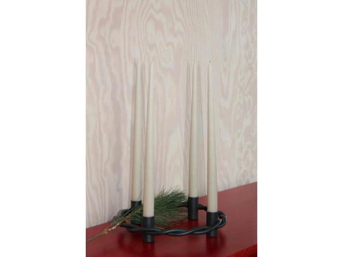 En sort metalstage med fire høje, cremefarvede stearinlys og en lille fyrretræsgren står på en rød flade mod en lys træmønstret væg.