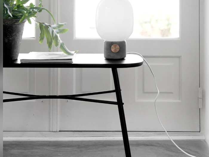 En moderne bordlampe med en hvid glasskærm står på et sort bord ved siden af en potteplante og en stak magasiner foran en hvid dør.