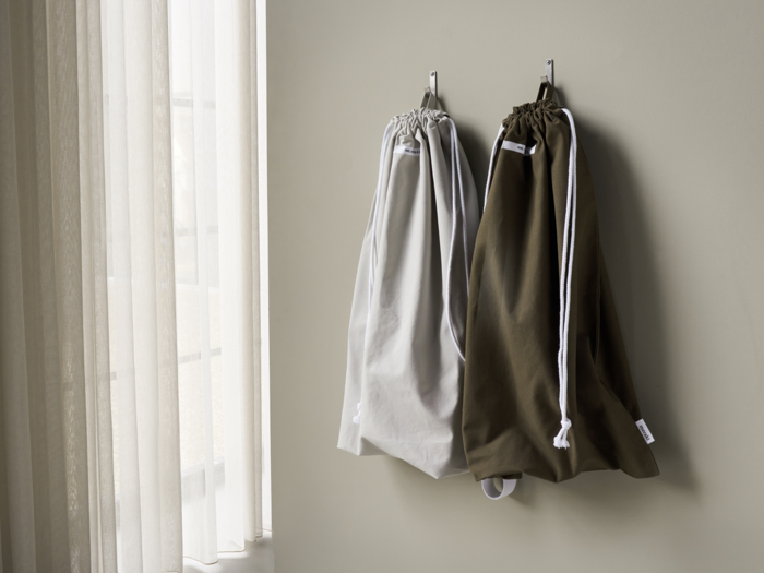 To poser med snor, en lysegrå og en mørk olivengrøn, hænger på kroge på en lys væg ved siden af et vindue med gennemsigtige gardiner.