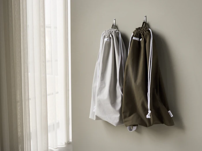 To poser med snor, en lysegrå og en mørk olivengrøn, hænger på kroge på en lys væg ved siden af et vindue med gennemsigtige gardiner.