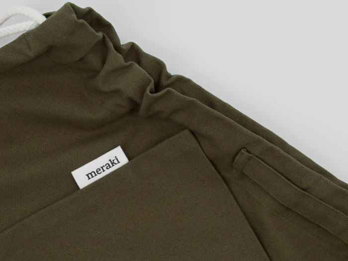 Nærbillede af en olivengrøn beklædningsgenstand med en lomme, hvid løbegang og et stofmærke, hvor der står "meraki".