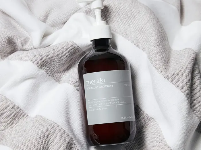 En flaske Meraki volumengivende balsam med pumpe står på en hvid- og gråstribet stofbaggrund.