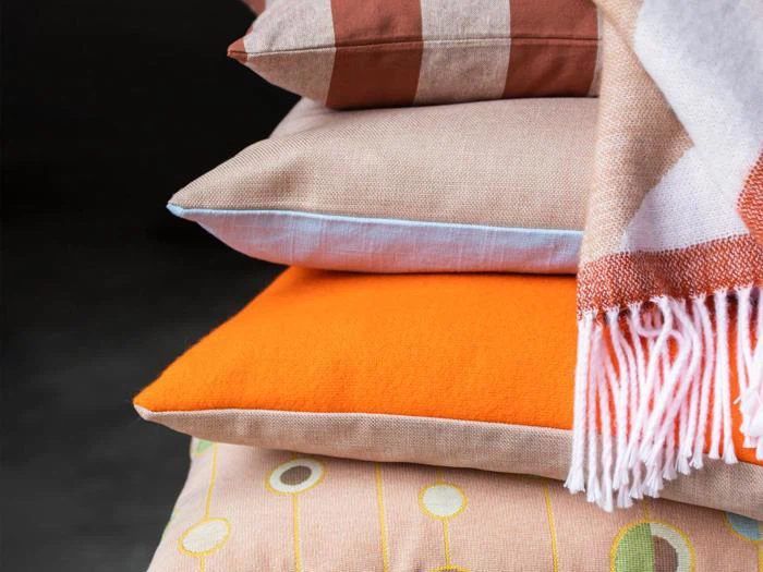 En stak farverige puder med forskellige teksturer og mønstre, toppet med et stribet tæppe, mod en mørk baggrund.