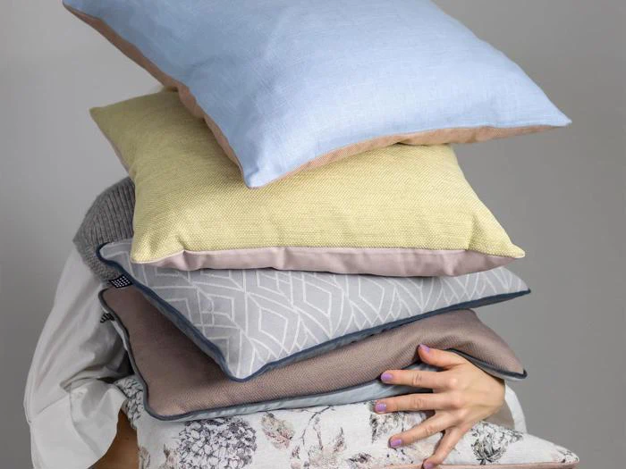 En person holder en stak med fem farverige puder, der dækker deres ansigt, med en lysegrå baggrund.