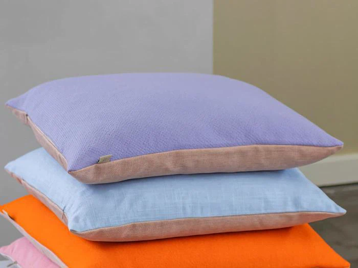 En stak med tre puder: den øverste pude er lavendel, den midterste er lyseblå, og den nederste er lys orange, alle med beige kanter, på en neutral baggrund.