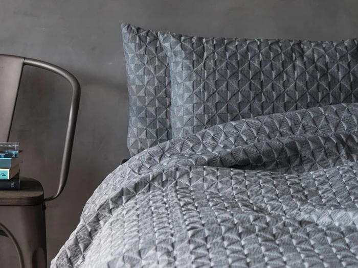 En pænt redt seng med et gråt quiltet tæppe og matchende puder med et geometrisk mønster ved siden af en metalstol med bøger på sædet.