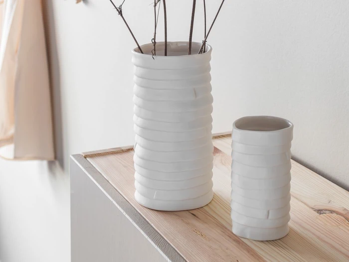 To hvide, strukturerede keramiske vaser står på en lys træoverflade. Den ene vase er høj og har små kviste indeni, mens den anden er lavere og tom. Baggrunden er minimalistisk og blødt oplyst.