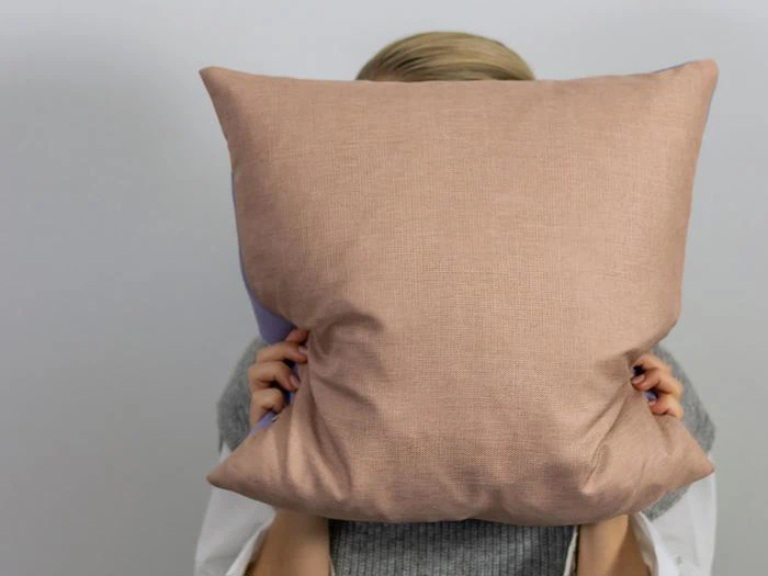 En person holder en stor, lysebrun pude foran ansigtet og skjuler sine ansigtstræk på en almindelig baggrund.