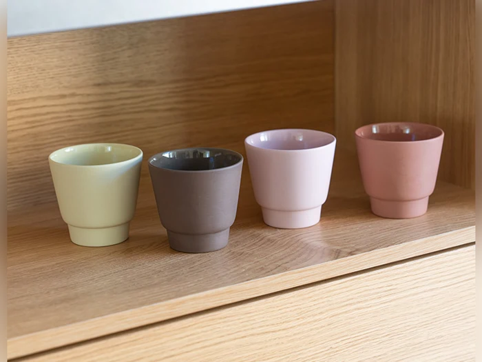 Fire små kopper i pastelfarver - lysegul, brun, lyserød og fersken - står pænt arrangeret på en lys træhylde.