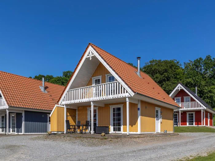 Et gult træhus med rødt tegltag, balkon og udendørs siddepladser under en klar blå himmel med træer i baggrunden.