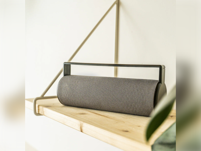 En moderne, cylindrisk grå højttaler med håndtag står på en lys træhylde mod en hvid væg med minimalistisk indretning.