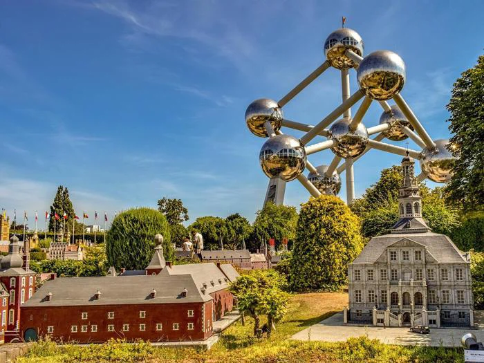 Atomium-monumentet tårner sig op bag miniaturebygninger og flag i en park på en solskinsdag, omgivet af grønt og blå himmel.