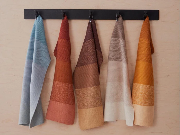 Fem farvede håndklæder i nuancer af blå, rød, brun, beige og orange hænger på sorte kroge mod en lys trævæg.