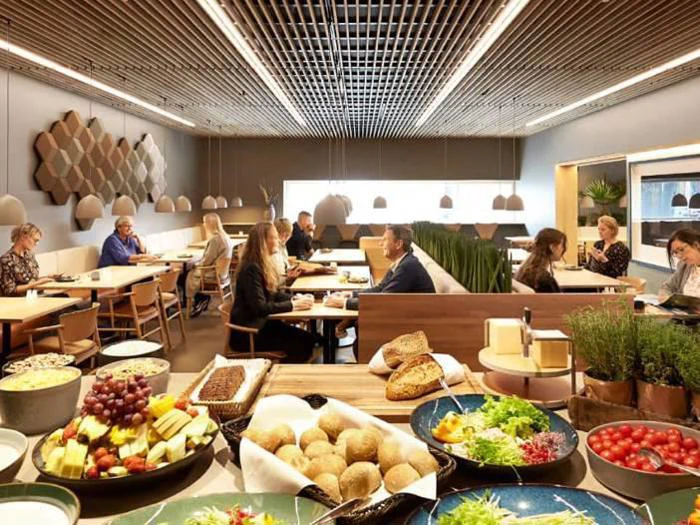 Moderne cafeteria med mennesker, der spiser ved borde; en buffet i forgrunden indeholder brød, frugt, ost og salat; varm belysning og moderne indretning.