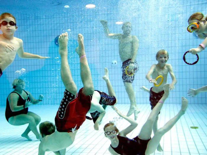 En gruppe børn og voksne iført badetøj og svømmebriller leger under vandet i en swimmingpool, omgivet af pool-legetøj og flydende ringe.