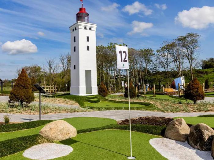 En minigolfbane med en hvid fyrtårnsstruktur, grønt kunstgræs, et flag med tallet 12, klipper og træer under en blå himmel med skyer.