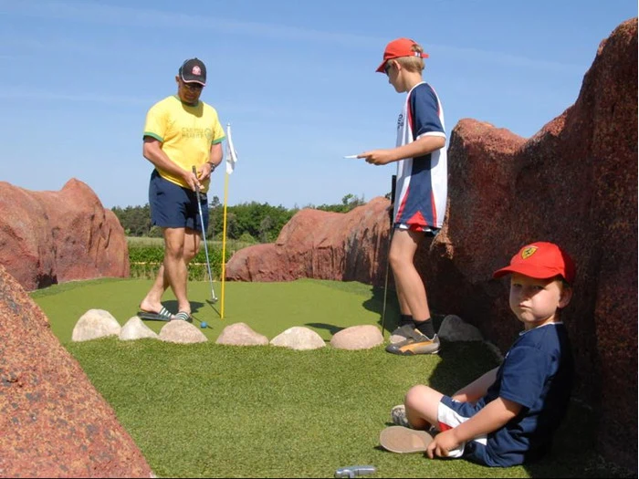 Tre personer er på en minigolfbane; en voksen spiller, et barn står med et scorekort, og et andet barn sidder på kunstgræsset ved siden af klipperne på en solrig dag.