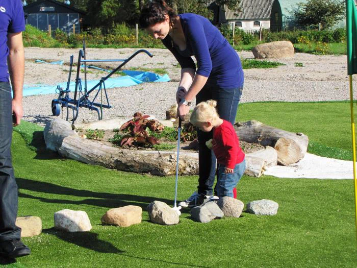 En kvinde hjælper et lille barn med at putte en golfbold på en minigolfbane med sten, der markerer hullet, og grønne områder i baggrunden.