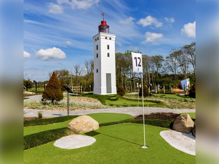 En minigolfbane med kunstgræs, klipper og et flag mærket med tallet 12, beliggende tæt på et højt, hvidt fyrtårn under en blå himmel med spredte skyer.