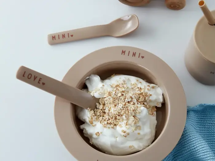 En beige skål mærket MINI fyldt med yoghurt og havregryn med en matchende ske mærket LOVE. En beige ske, en kop med sugerør og en blå klud ligger i nærheden på en hvid flade.