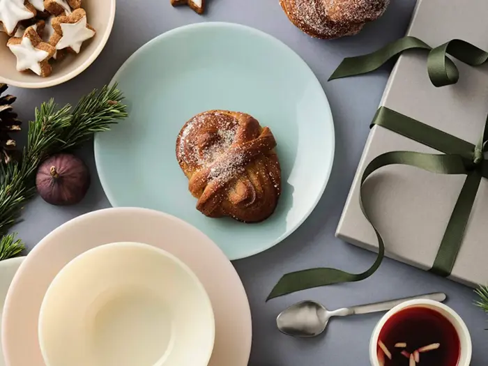 Et frostet wienerbrød på en blå tallerken, en skål, en kop te, småkager, grønne planter og en grå gaveæske med et grønt bånd står på et bord.