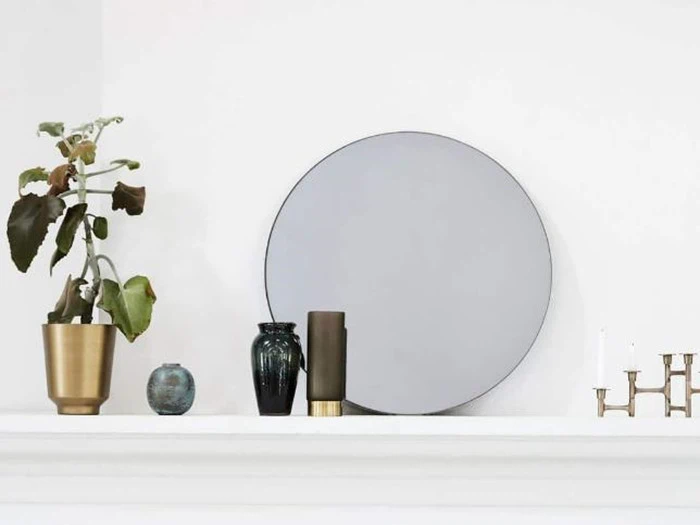 Et rundt spejl, en potteplante, vaser og en lysestage er anbragt på en hvid kaminhylde mod en almindelig væg.