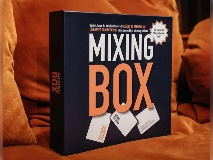 En brætspilsæske med titlen Mixing Box ligger på en orange sofa. Æsken har fed hvid og orange tekst på en mørk baggrund, og kortene er illustreret i bunden.