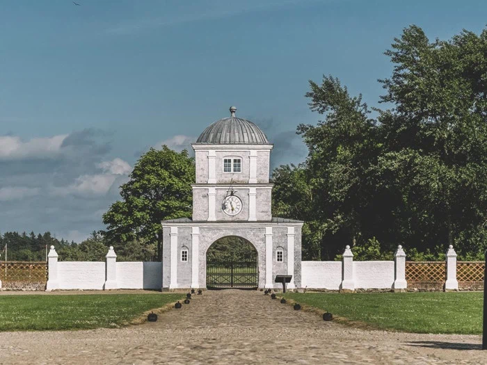 En hvid stenport med et ur og en buet indgang står foran en grøn græsplæne, træer og en blå himmel.