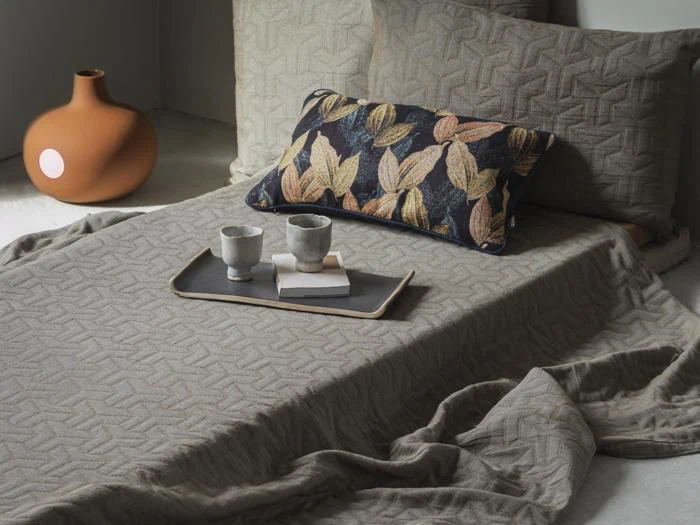 En hyggelig seng med mønstret gråt sengetøj, en sort pude med bladmotiver og en bakke med to kopper og en bog. En orange vase står på gulvet i nærheden.