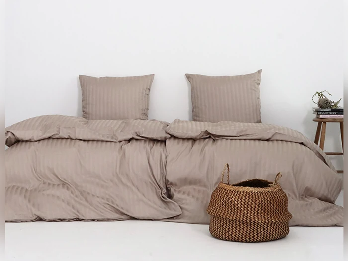 En pænt redt seng med taupefarvet sengetøj og to puder, en flettet kurv på gulvet og et lille sidebord med en pyntegenstand og en bog mod en hvid væg.